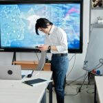 A man standing in an office checks his smartphone with a digital screen displaying AI graphics.
