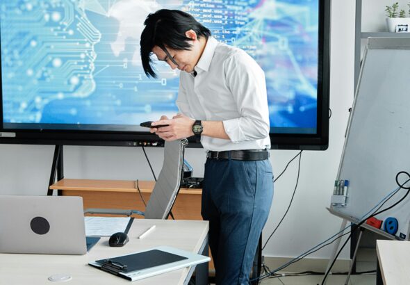 A man standing in an office checks his smartphone with a digital screen displaying AI graphics.